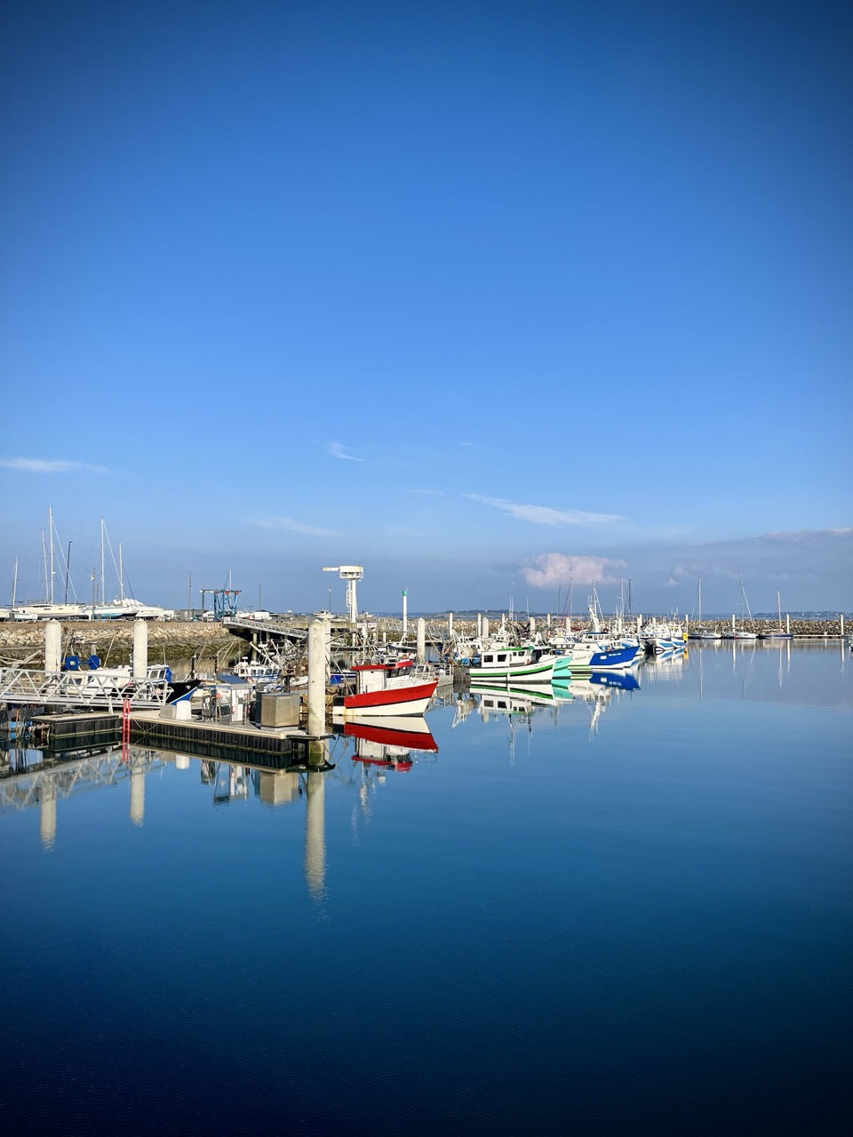 Le port de Saint-Cast, on y voit des bateaux de pecheurs aux couleurs vives, bleu, rouge, dans une mer bleue et un ciel bleu. Lumière douce de fin de journée